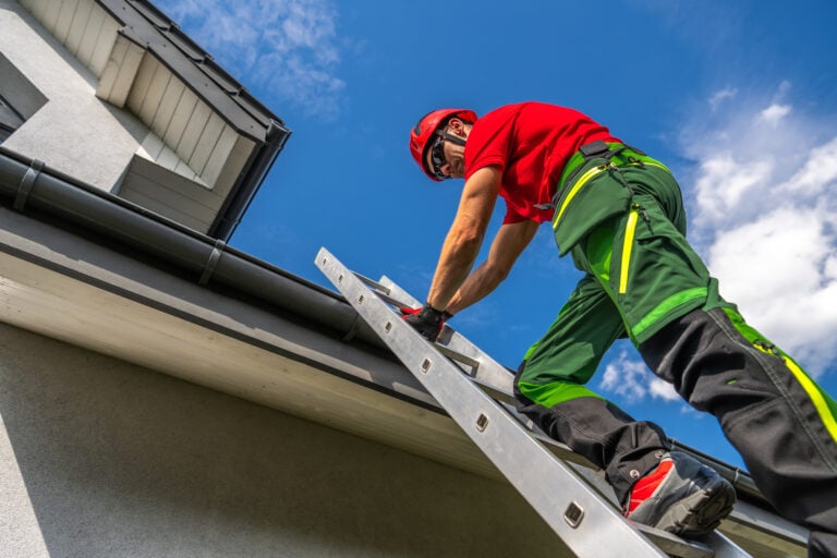 A maintenance worker in safety gear ascends a ladder to inspect and repair a roof gutter while the sun shines brightly overhead.