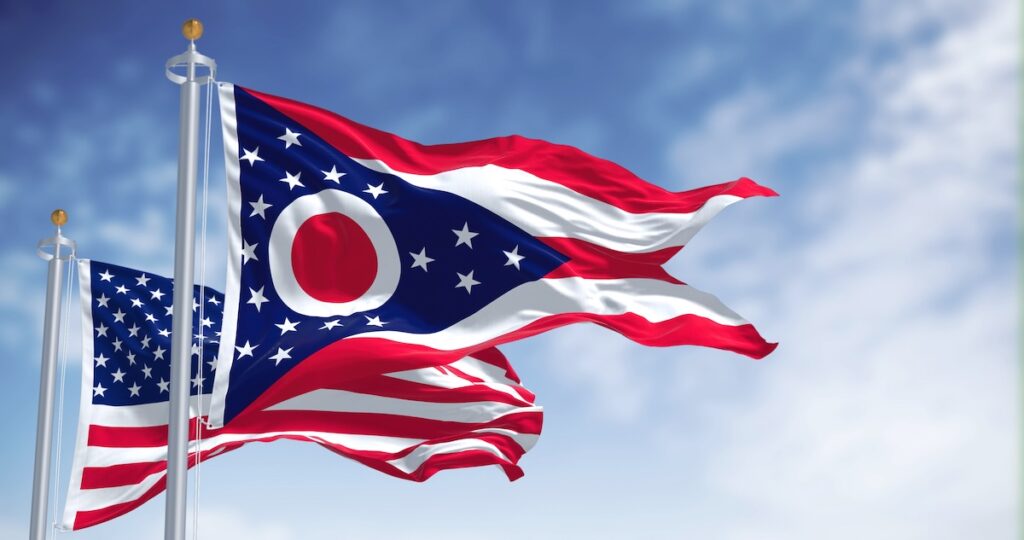 The Ohio state flag waving along with the national flag of the United States of America. In the background there is a clear sky. Ohio is a state in the Midwestern region of the United States